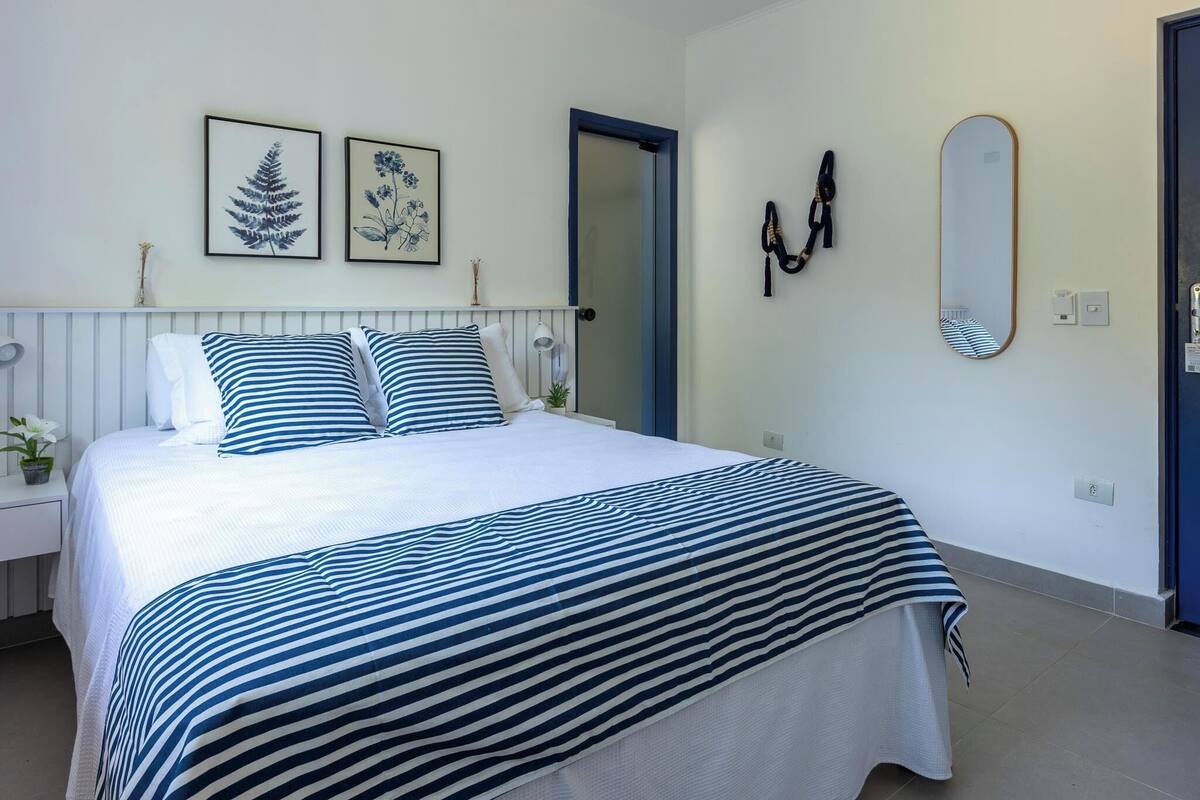 A neatly made bed with striped bedding, a bedside table with a lamp, and two framed botanical prints on the wall.