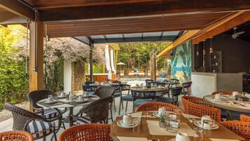 Un área de comedor al aire libre con sillas y mesas de mimbre, una piscina al fondo y un mural en la pared.