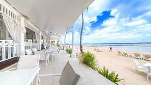 Une terrasse au bord de la plage, avec des chaises et des tables blanches, un espace salon couvert et une vue sur la plage et l’océan.