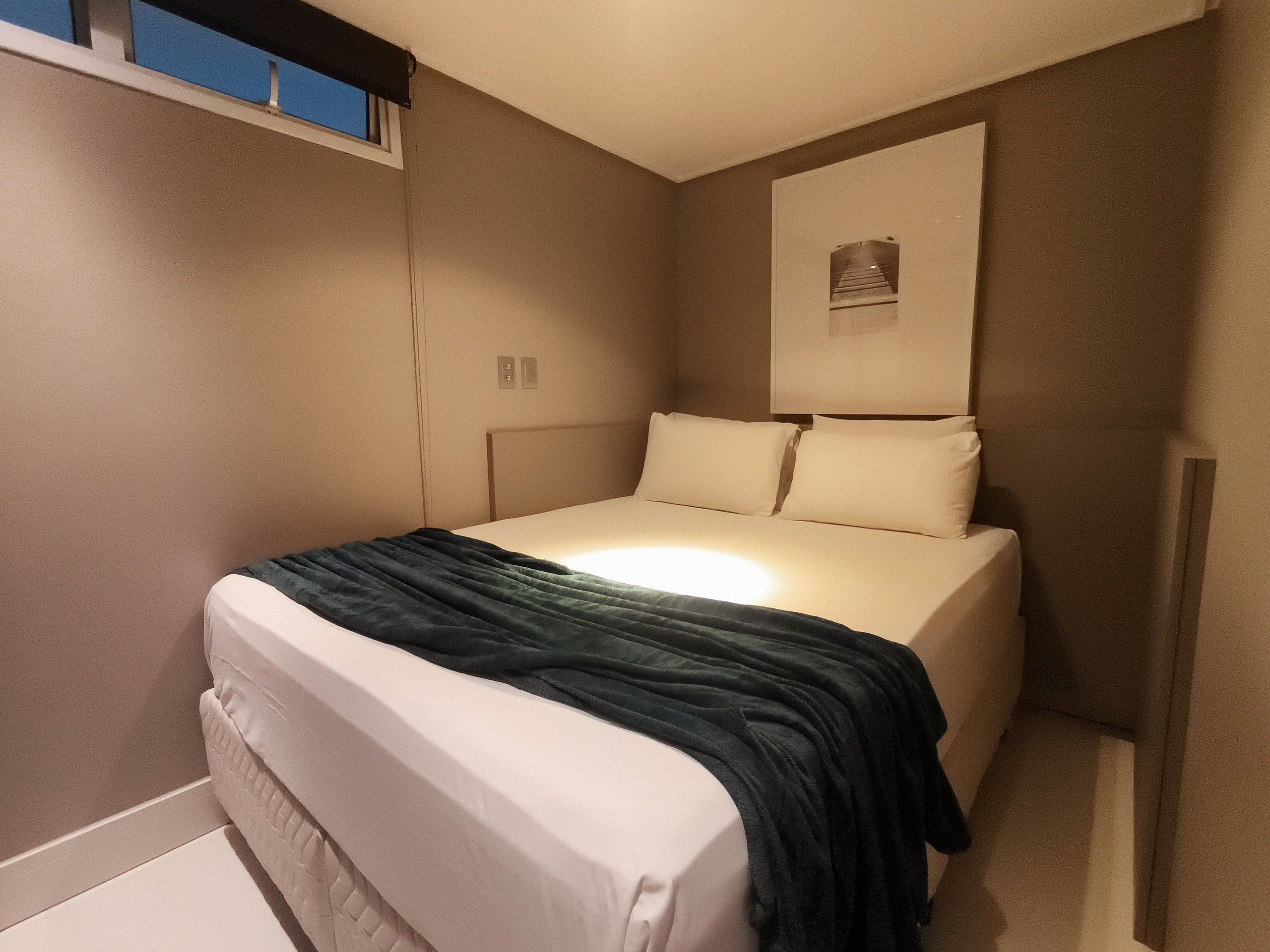 Una cama individual con una manta oscura, cabecera e iluminación empotrada en la pared.