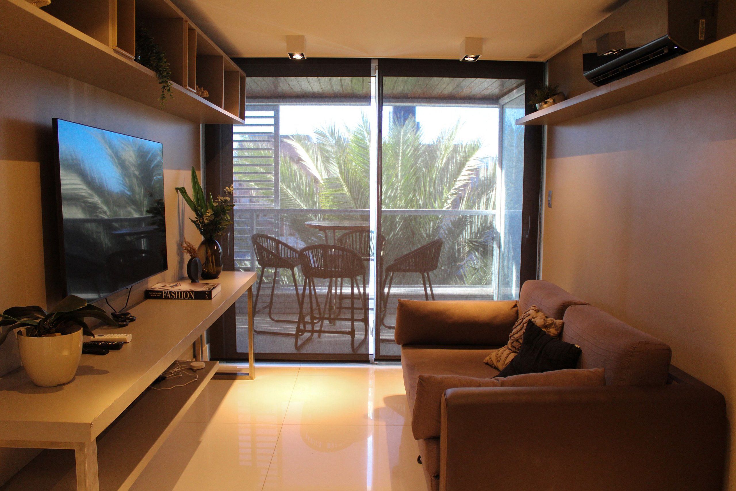 A modern living room with a flat-screen TV, a sofa, a coffee table, and a sliding glass door leading to an outdoor patio with chairs.
