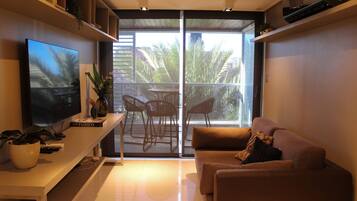 A modern living room with a flat-screen TV, a sofa, a coffee table, and a sliding glass door leading to an outdoor patio with chairs.