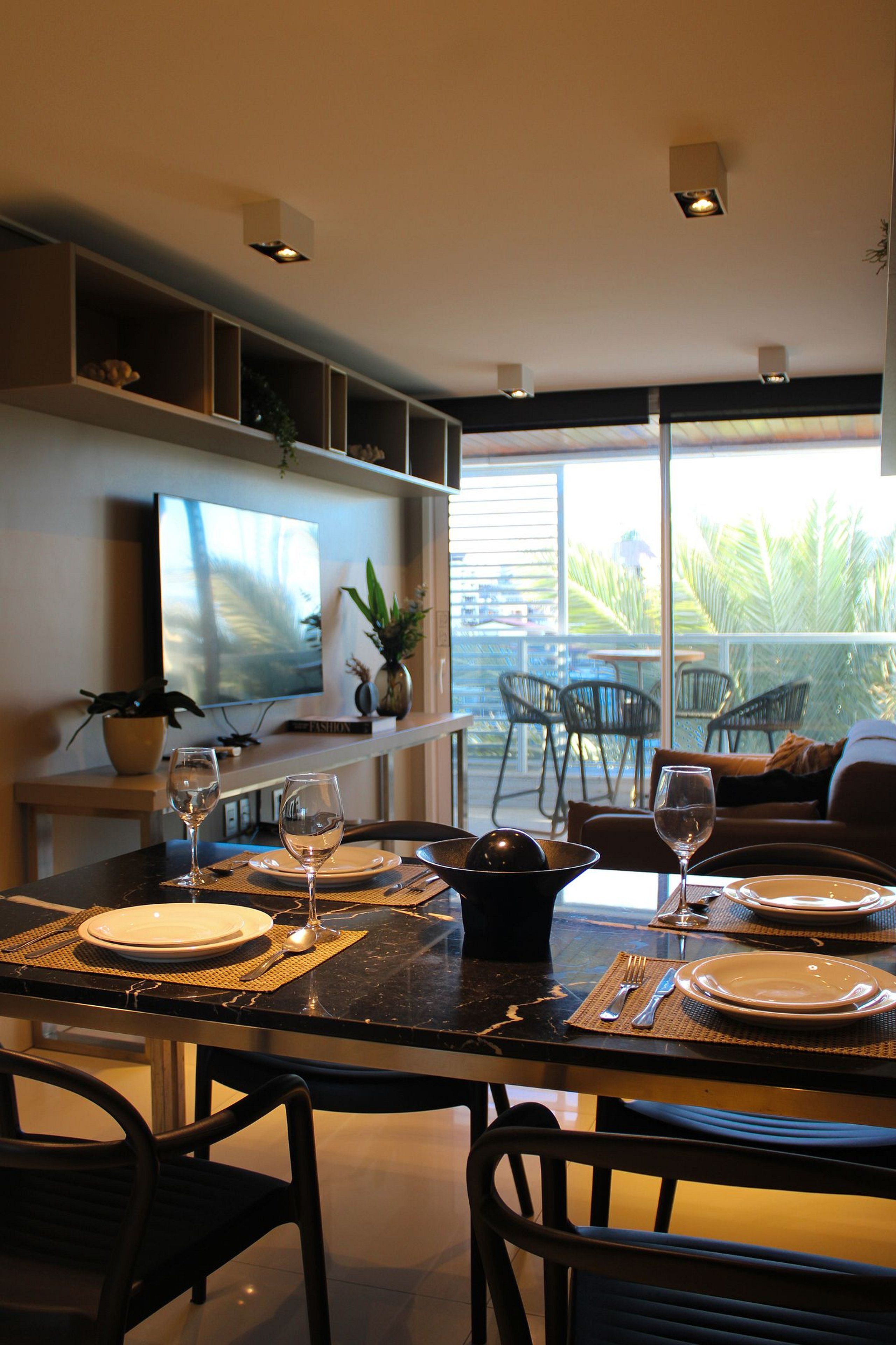 A dining area with a table set for two, a television mounted on the wall, and a view of the outdoors.