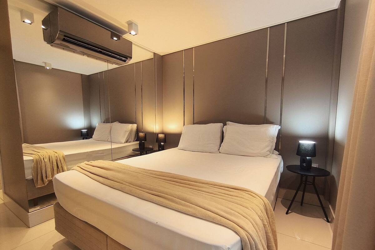 A modern hotel room with a large bed, bedside tables, a mirror, and an air conditioning unit.