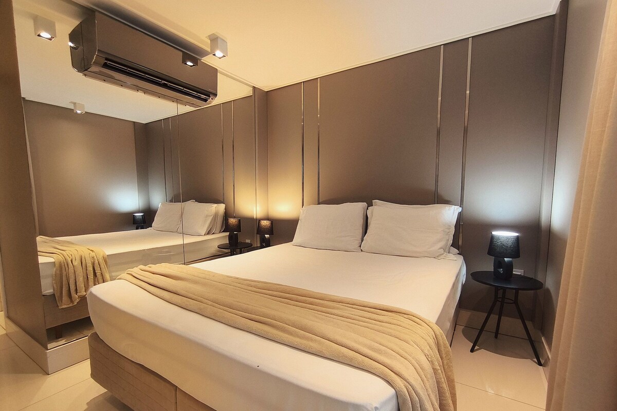 A modern hotel room with a large bed, bedside tables, a mirror, and an air conditioning unit.