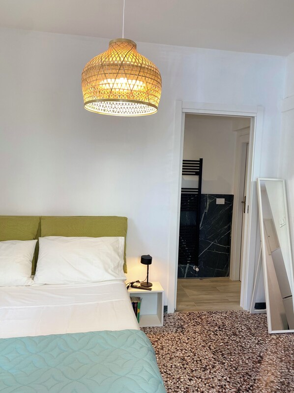 Un dormitorio con cama, lámpara de noche, lámpara colgante y puerta que da a un baño.