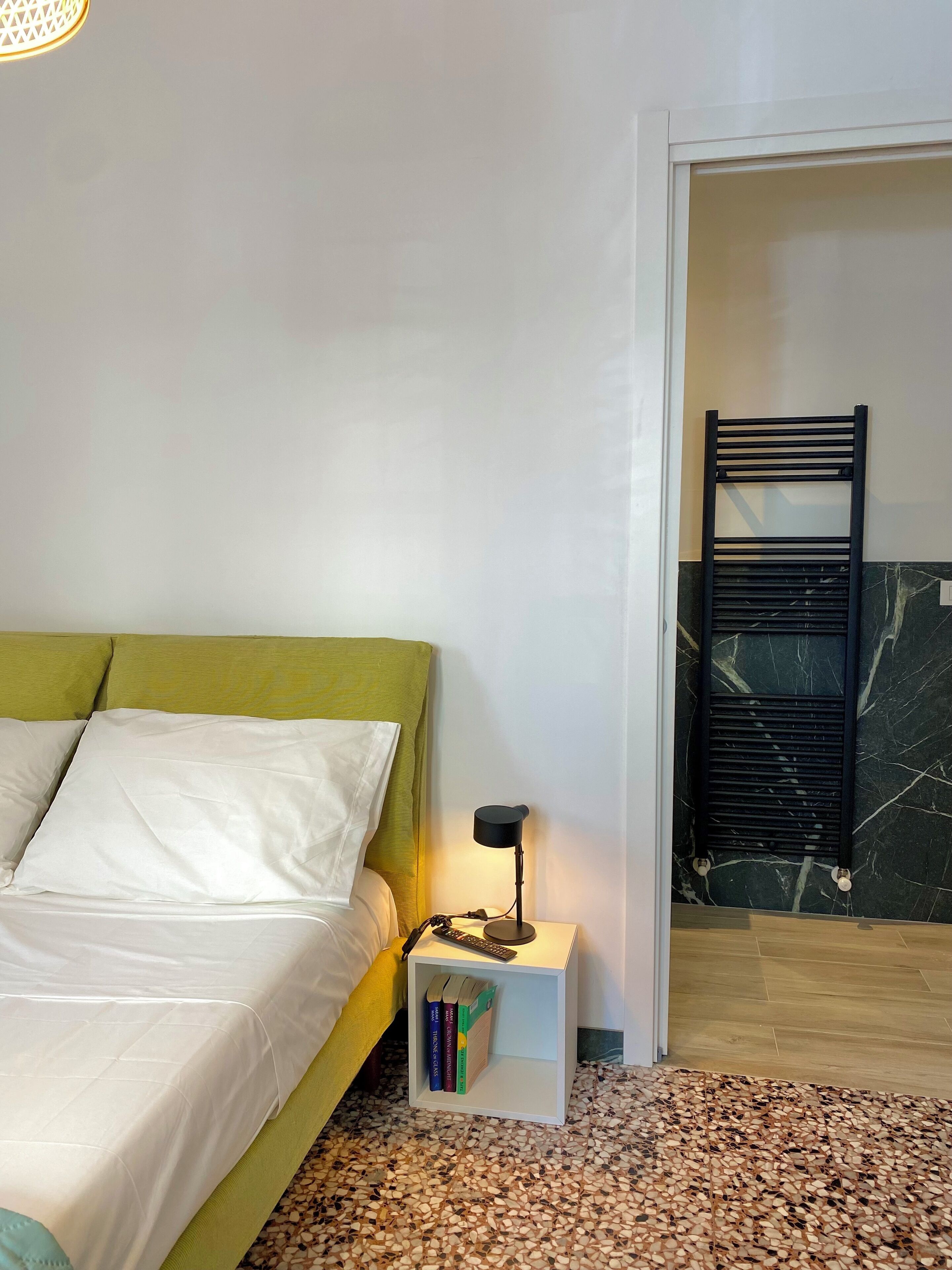Una camera da letto con un letto, un comodino con dei libri, un tavolino con una lampada e un termosifone.