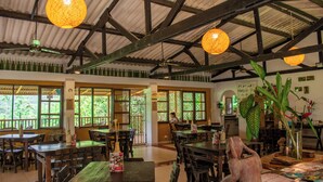 A restaurant with wooden tables and chairs, large windows, and hanging lights. - HOTEL MINCA LA CASONA (Santa Marta)