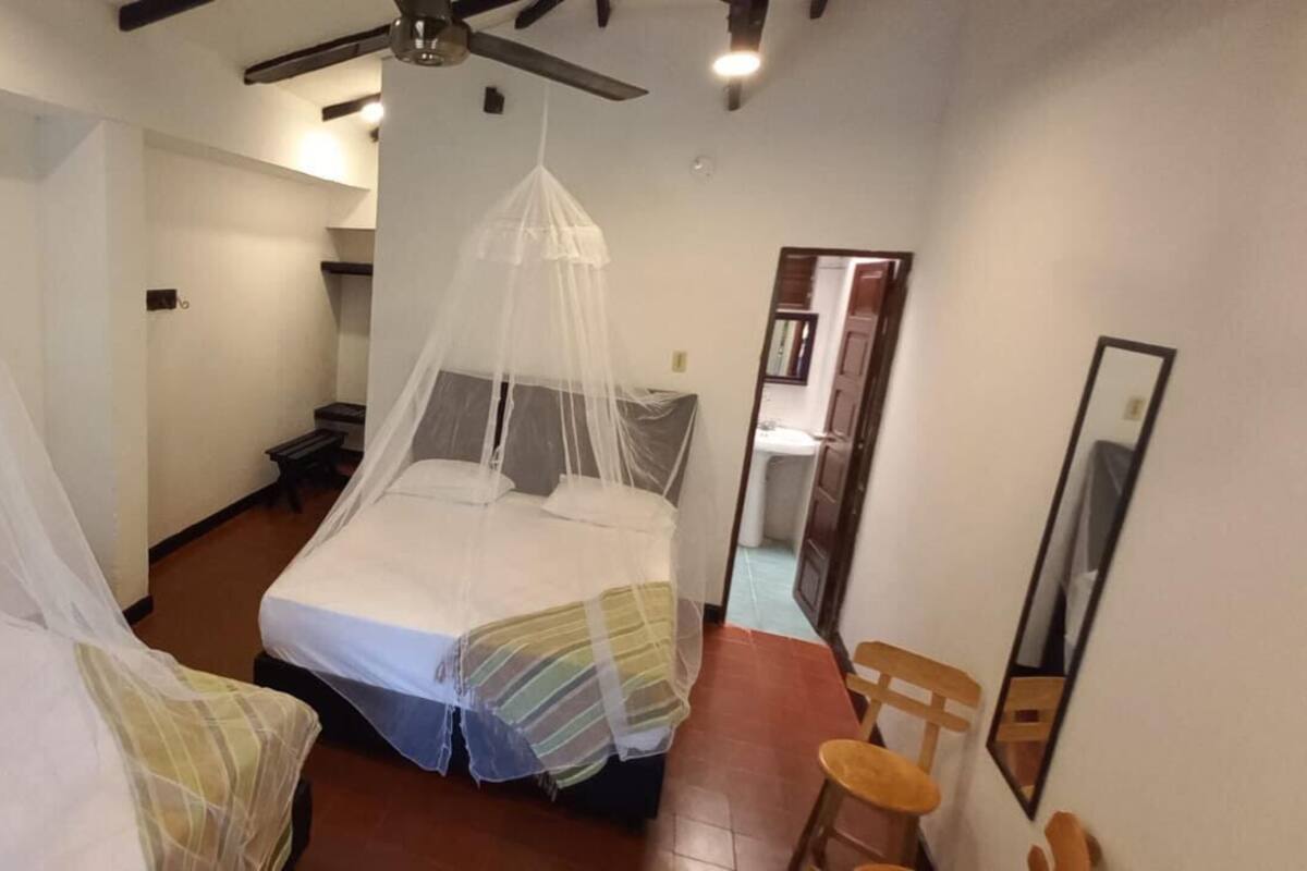 Habitación de hotel con dos camas, cada una con dosel, una mesita de noche de madera, un espejo y un ventilador de techo.
