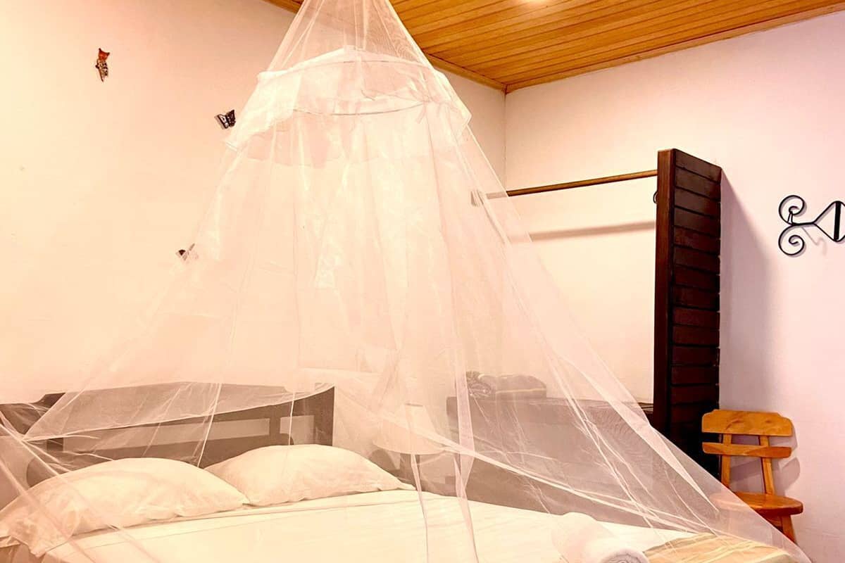 Un dormitorio con una cama cubierta por una mosquitera, techo de madera, estante en la pared y una pequeña silla de madera.
