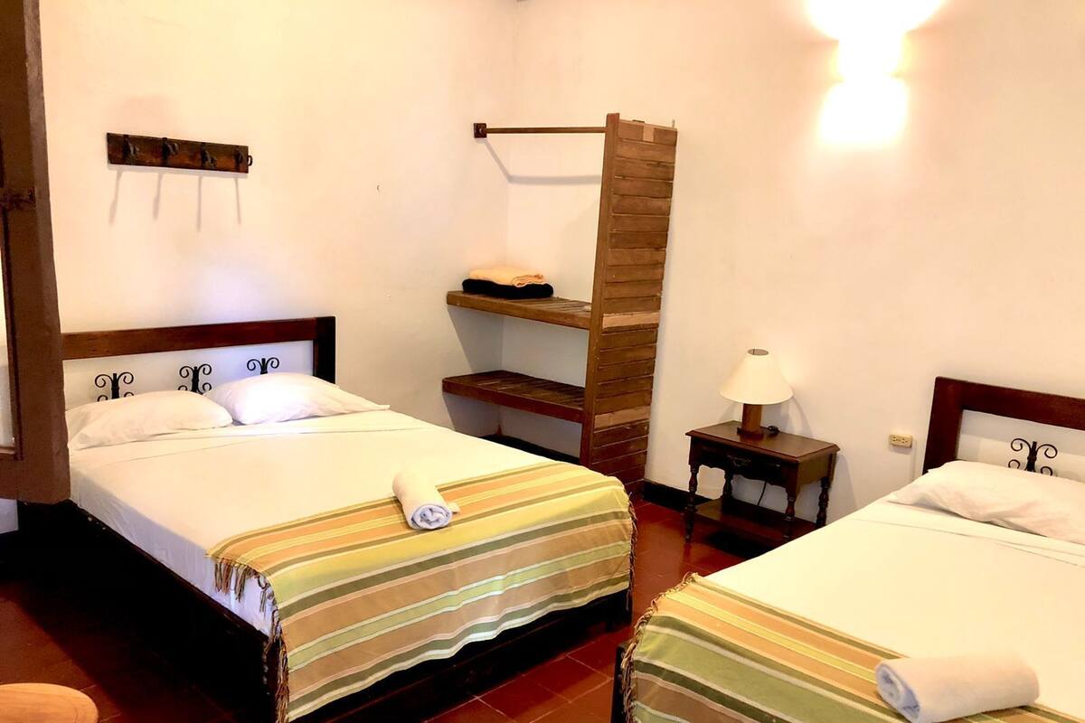 Habitación de hotel con dos camas, un estante de madera, un perchero y una pequeña mesa con una lámpara.