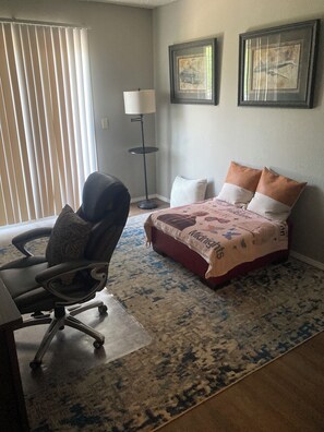 Une chambre à coucher comprenant un lit, un bureau, une chaise, une lampe sur pied et deux tableaux encadrés accrochés au mur.