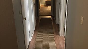 Un couloir doté d’un plancher en bois, d’un tapis à motifs et d’une porte donnant sur une pièce où se trouve une lampe.