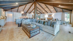 Een ruime loft met een hoge plafond, zichtbare houten balken, een grote zithoek met een bank en salontafel, een keuken met witte kastjes en een uitzicht door een groot raam.
