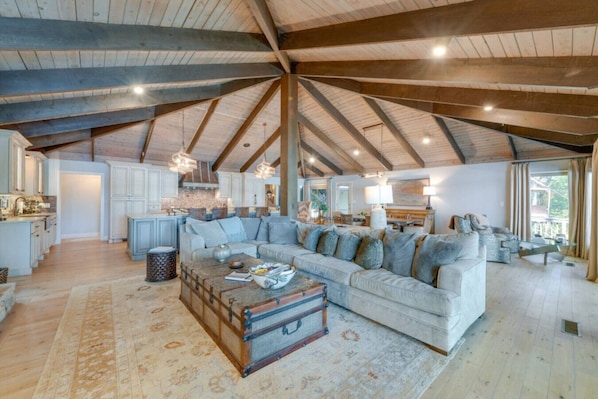 A spacious loft with a high ceiling, exposed wooden beams, a large living area with a sofa and a coffee table, a kitchen with white cabinets, and a view through a large window.
