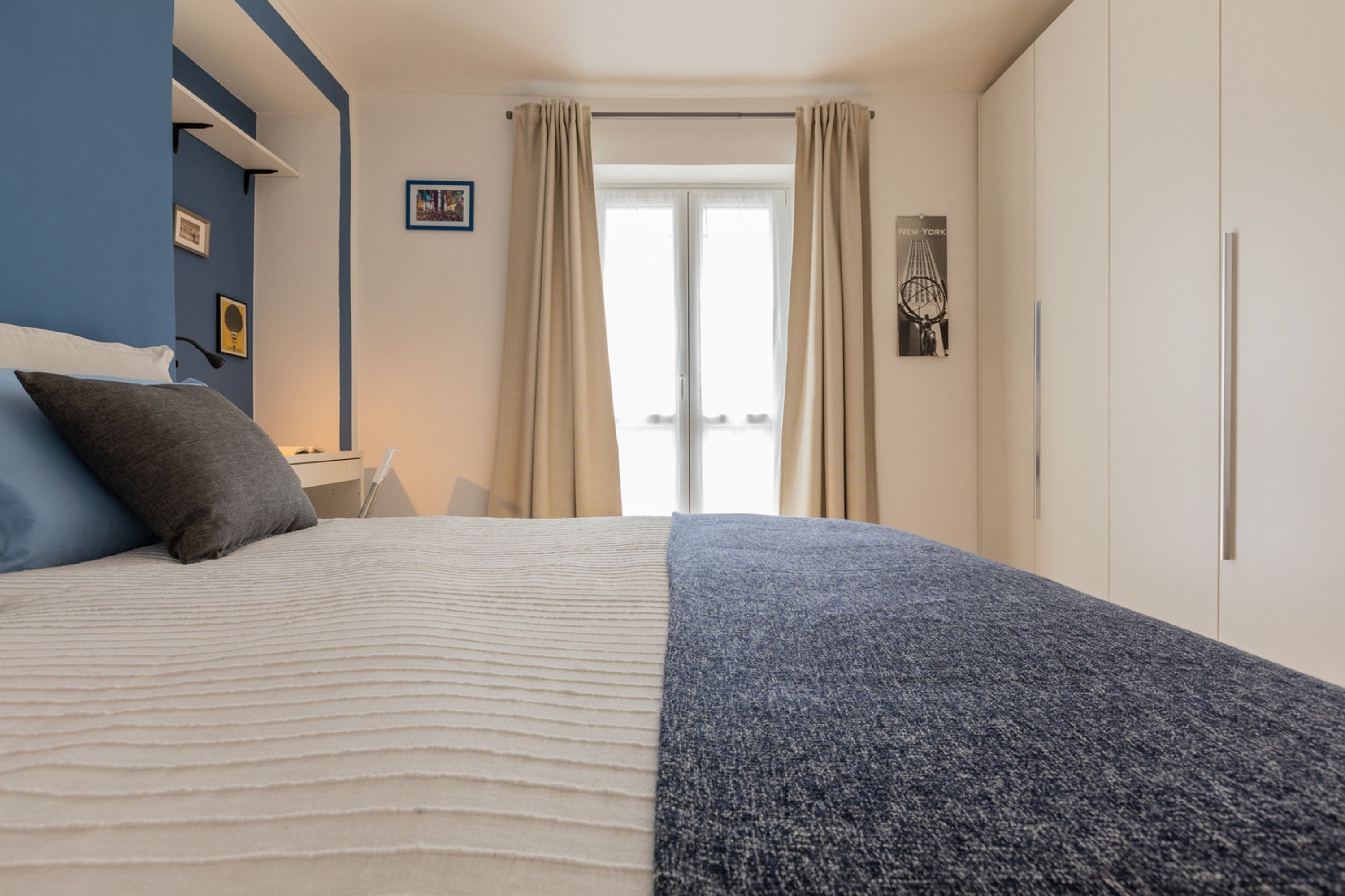 A neatly made bed with a white and blue blanket, a blue wall, and a window with curtains.