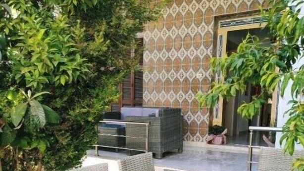 An outdoor patio with wicker furniture and a glass table, surrounded by greenery and a patterned wall.