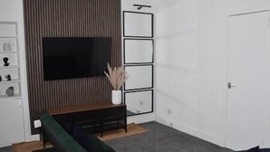 Un salon avec un téléviseur à écran plat fixé au mur, un canapé de couleur sombre et une petite table blanche sur laquelle est posé un vase contenant des plantes séchées.