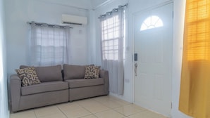 Una stanza con un divano color beige, due cuscini decorati, una porta bianca e una finestra con tenda.