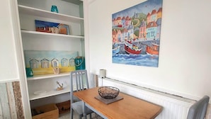 A dining area with a wooden table and grey upholstered chairs, a white radiator below, and a colorful painting of a harbor scene on the wall. - Cosy light and spacious apartment in the heart of Stratton  (Stratton)