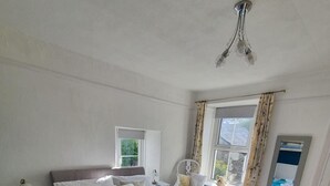 A bedroom with a bed, a nightstand, a chair, a window with curtains, and a ceiling light fixture. - Cosy light and spacious apartment in the heart of Stratton  (Stratton)