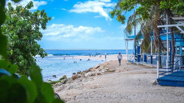 Une plage bordée d’un rivage rocheux, un ciel bleu dégagé et quelques personnes dans l’eau.