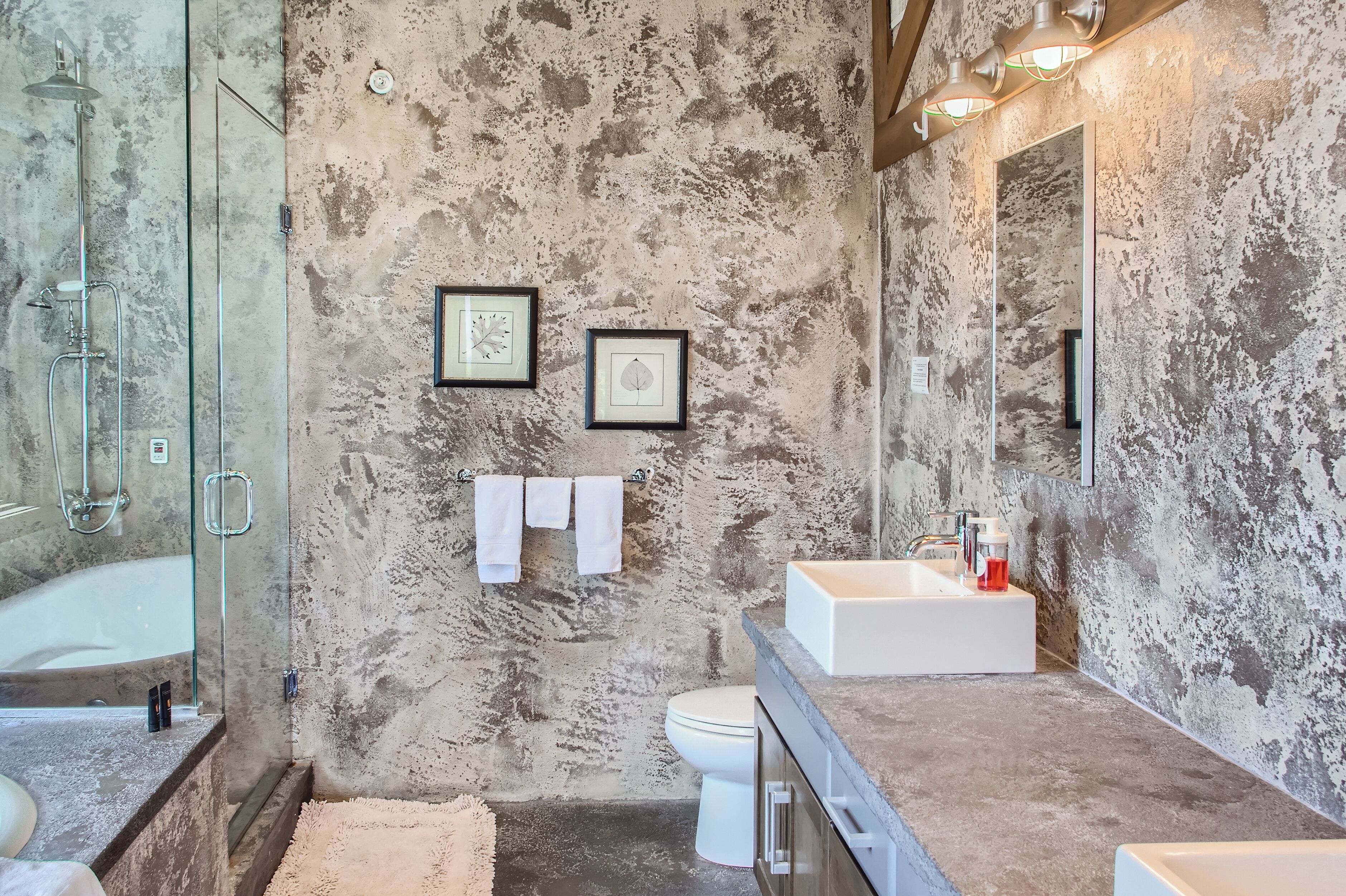 Un baño con pared de concreto, ducha con cerramiento de vidrio, inodoro blanco y lavamanos blanco con encimera.