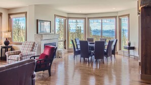 A spacious living room with a fireplace, large windows offering mountain views, and a dining area with modern furniture. - The Timbers by Okanagan Premier (Beaverdell)