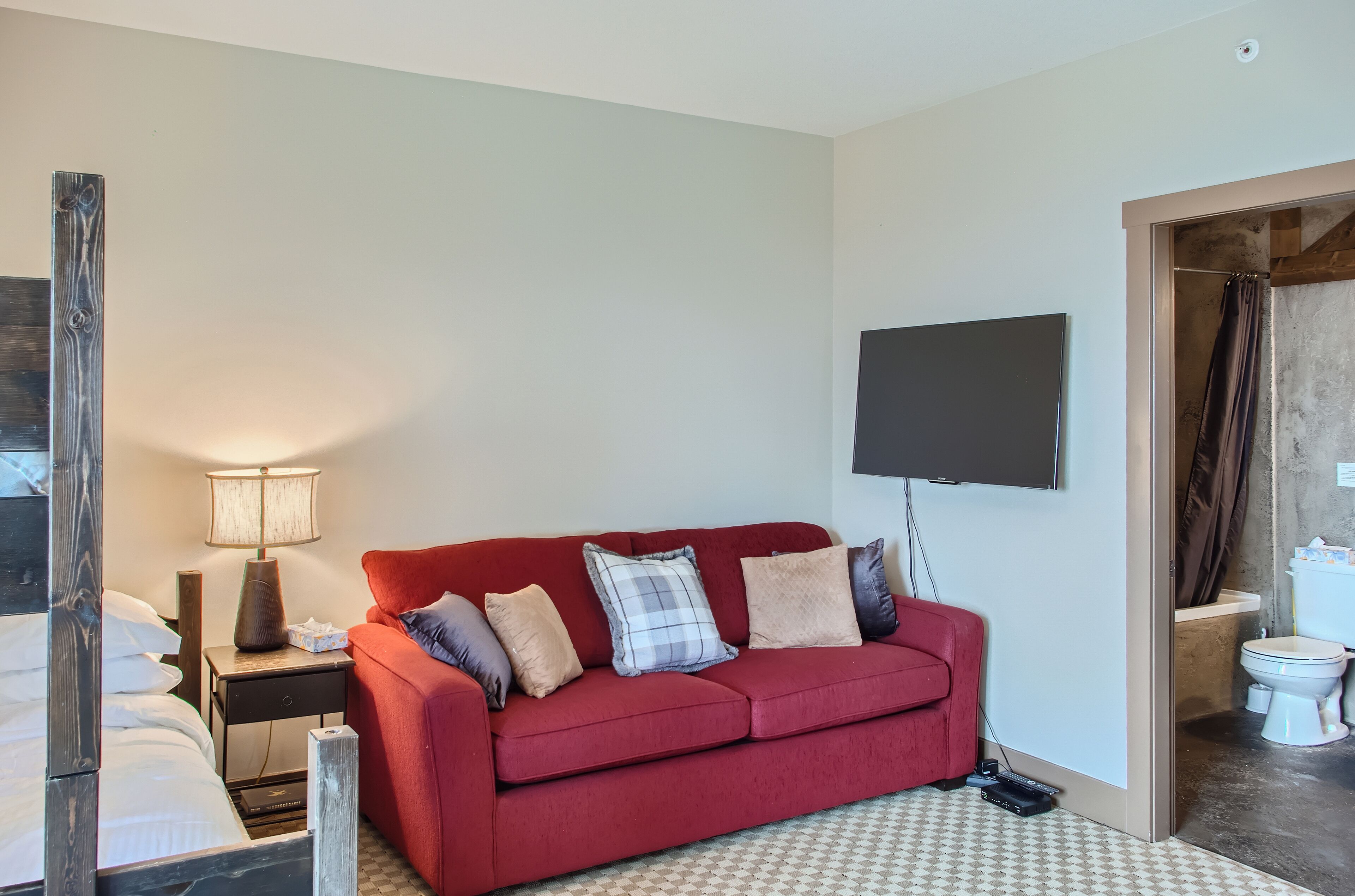 A hotel room with a red sofa, a flat-screen TV mounted on the wall, a bedside table with a lamp, and a bathroom visible through an open door.