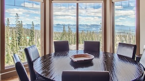 A dining area with a large round table, four chairs, and a view of a forest and mountains through large windows. - The Timbers by Okanagan Premier (Beaverdell)