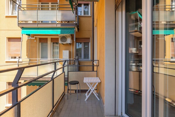 A balcony with a small table and chair, a window with a view of another building, and a green awning. - Appartamento Ibisco by Wonderful Italy (Bologna)