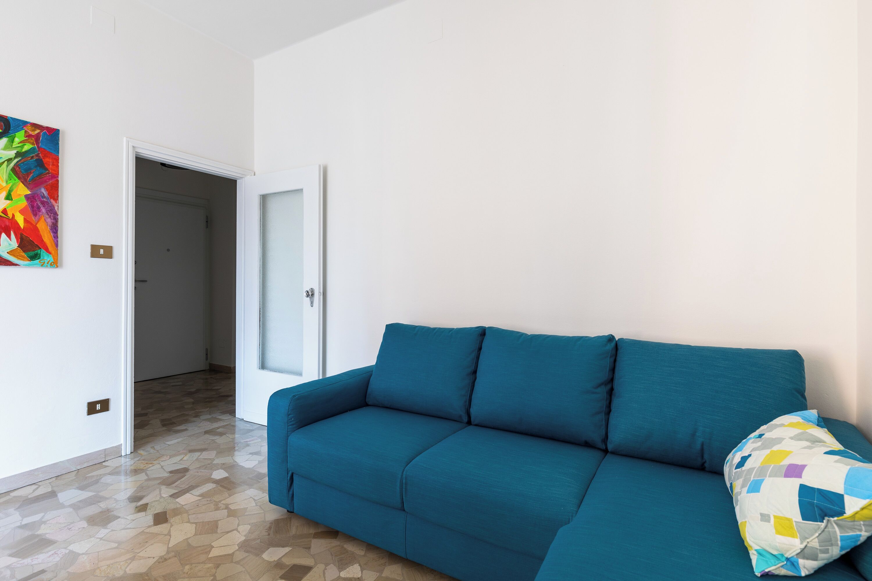 Un canapé de couleur turquoise agrémenté d’un coussin coloré, un sol carrelé et une porte donnant sur une autre pièce.