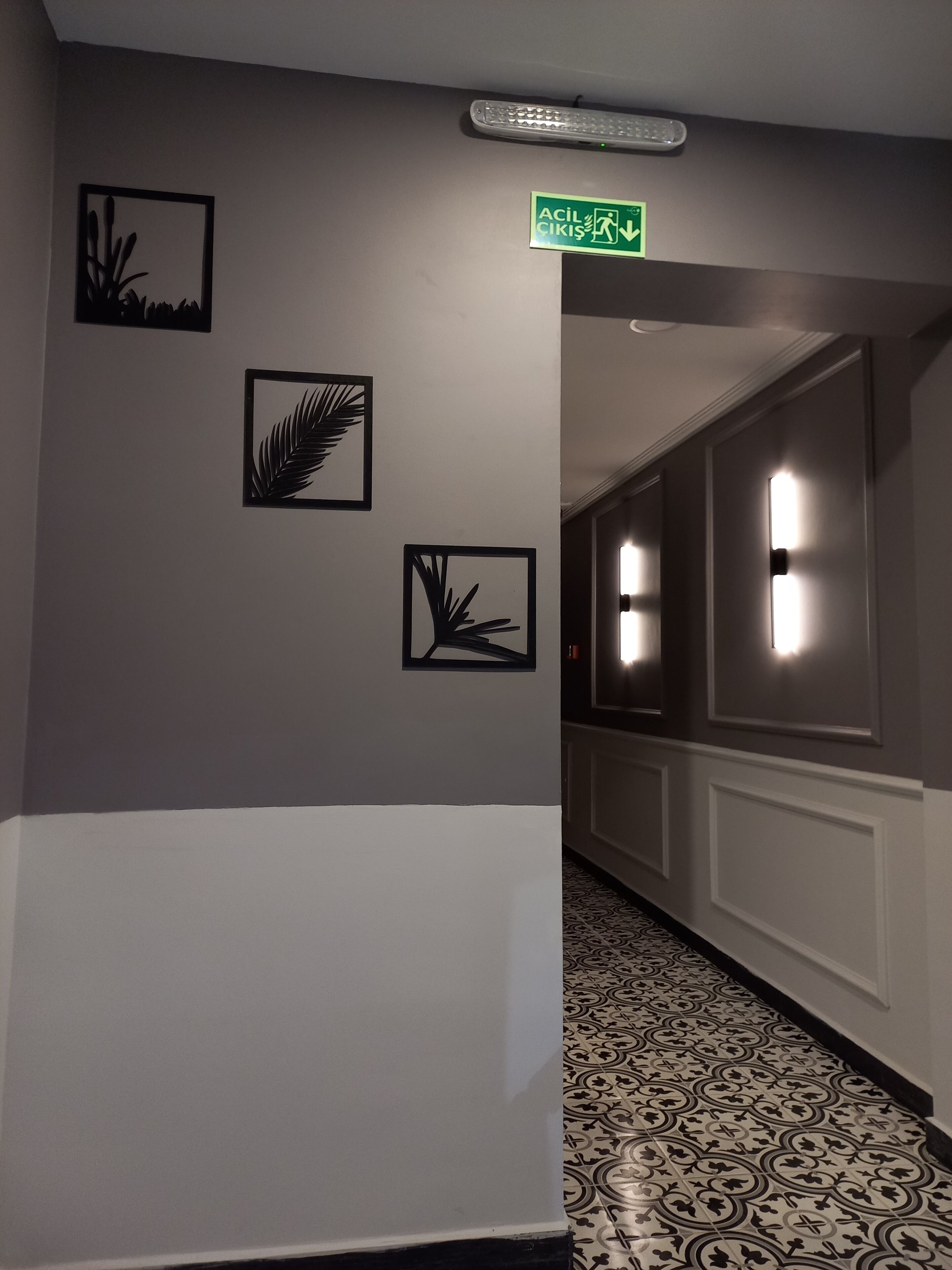 A hallway with patterned floor tiles, a green emergency exit sign, and three black wall art pieces.