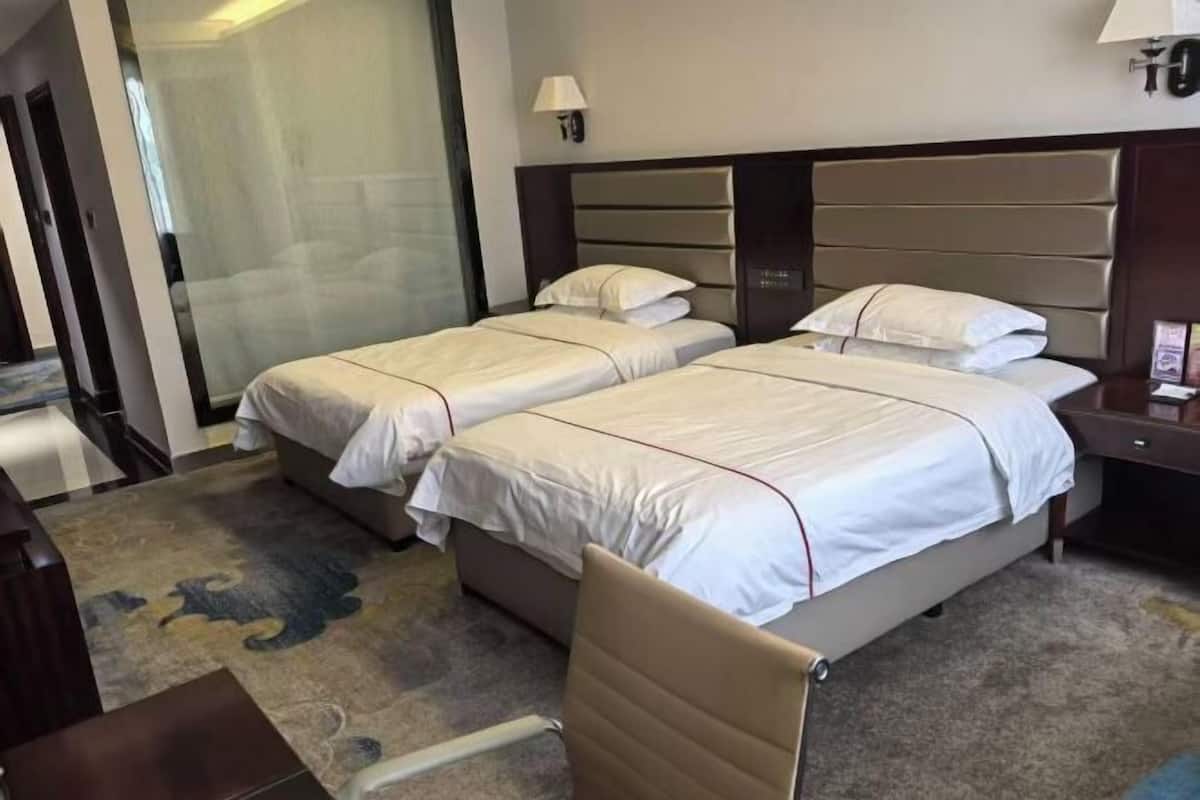 A hotel room with two beds, a desk, a chair, a lamp, and a glass partition.