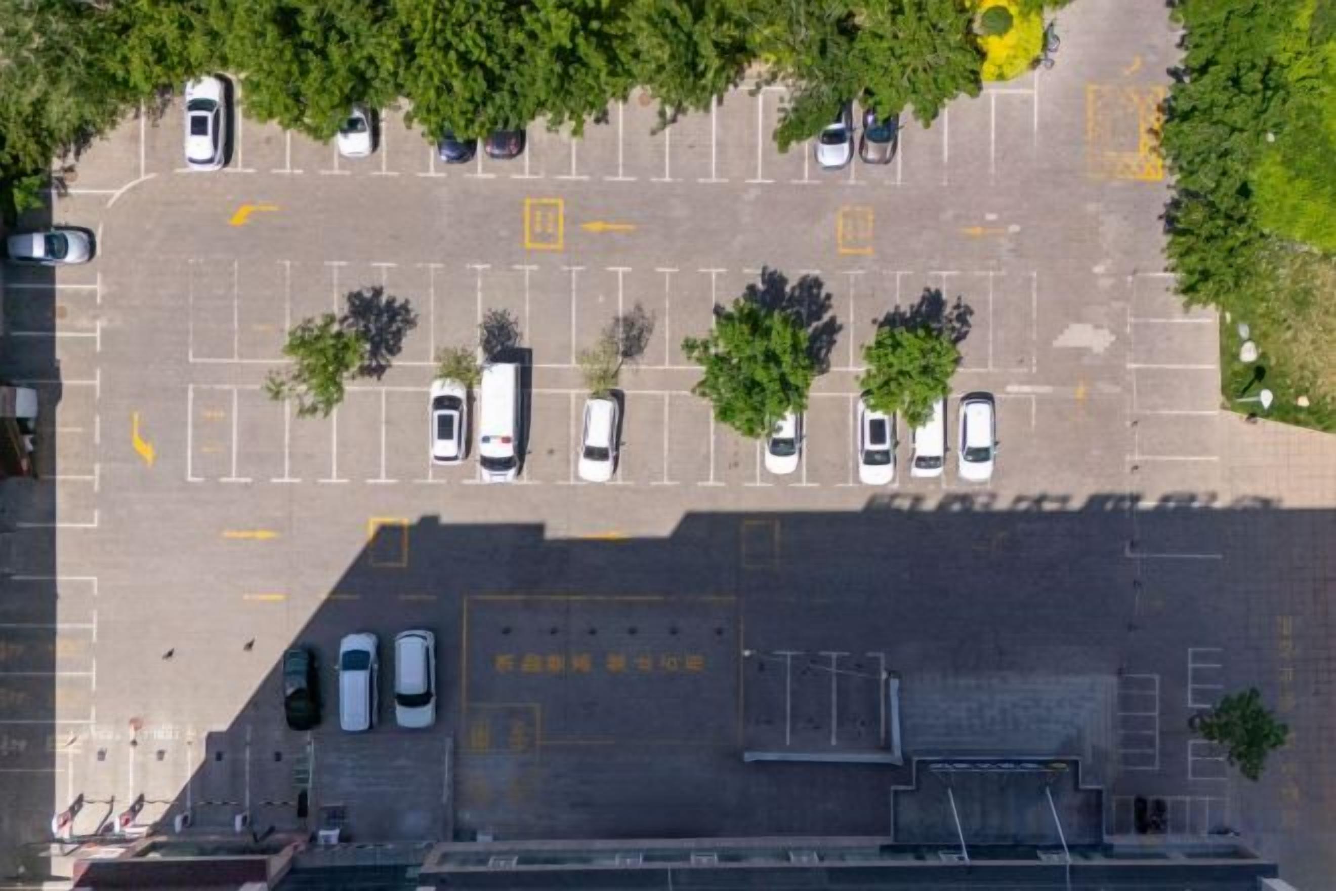 An aerial view of a parking lot with multiple cars, trees, and marked parking spaces.