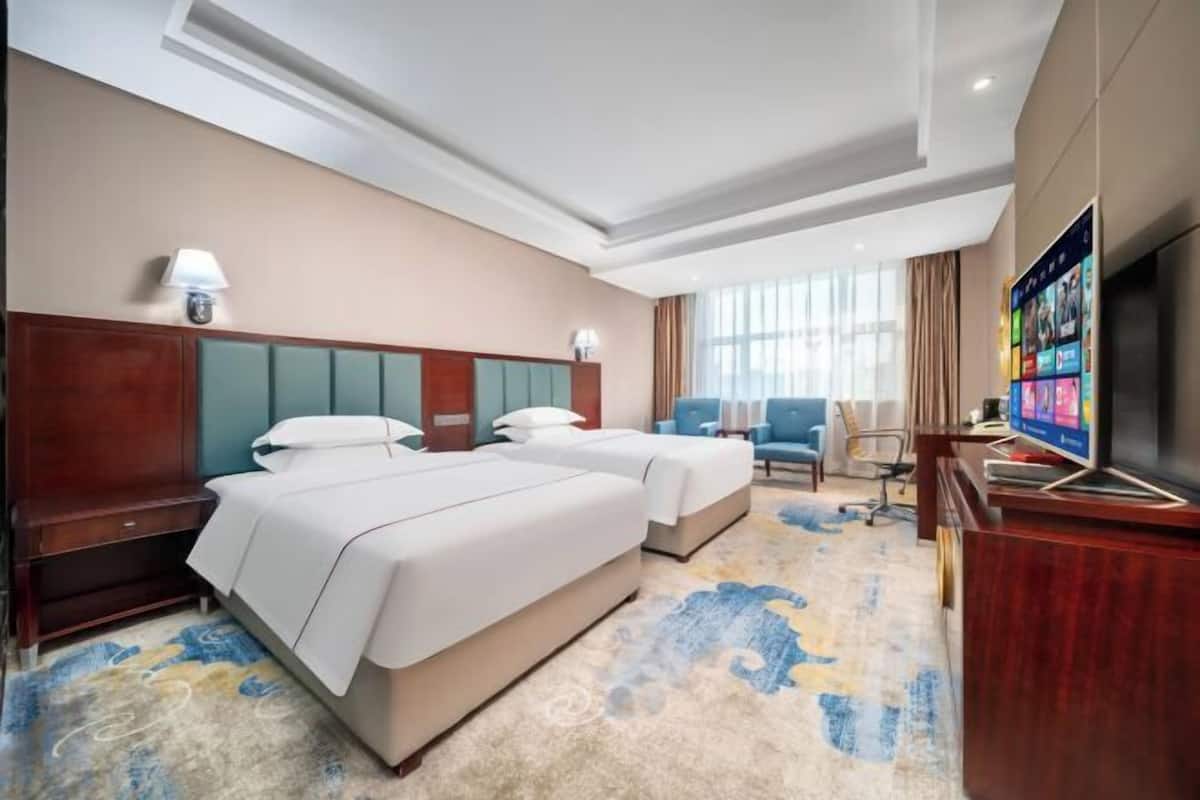 A hotel room with two beds, a TV, a desk, and a large window.