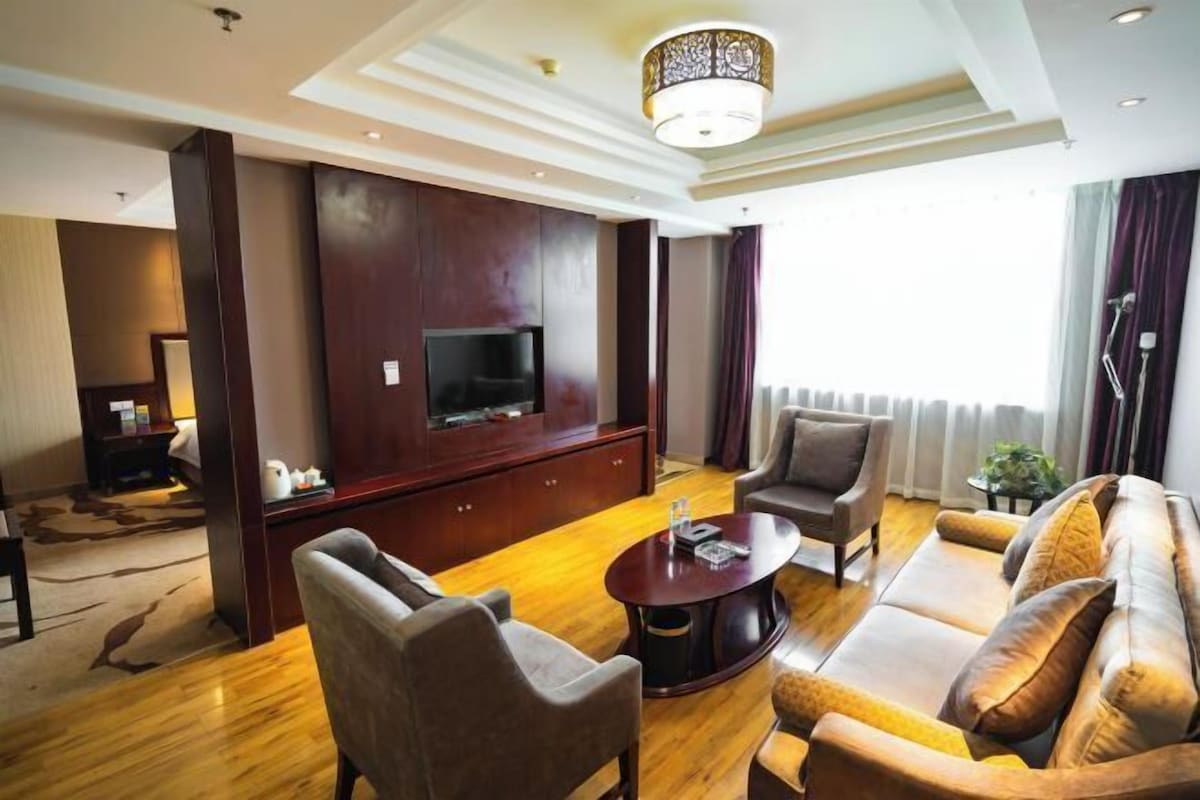 A modern living room with a flat-screen TV, leather sofas, a coffee table, and a large window with curtains.
