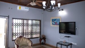 A room with a ceiling fan, a mounted TV, a window with metal bars, a framed picture, and a vintage chair. - Joyful Haven apartments (Nkoranza)