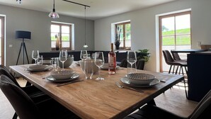 A dining area with a wooden table set for six, surrounded by chairs. There are two pendant lights above the table, and a floor lamp is visible to the left. The room has large windows with a view of greenery outside.