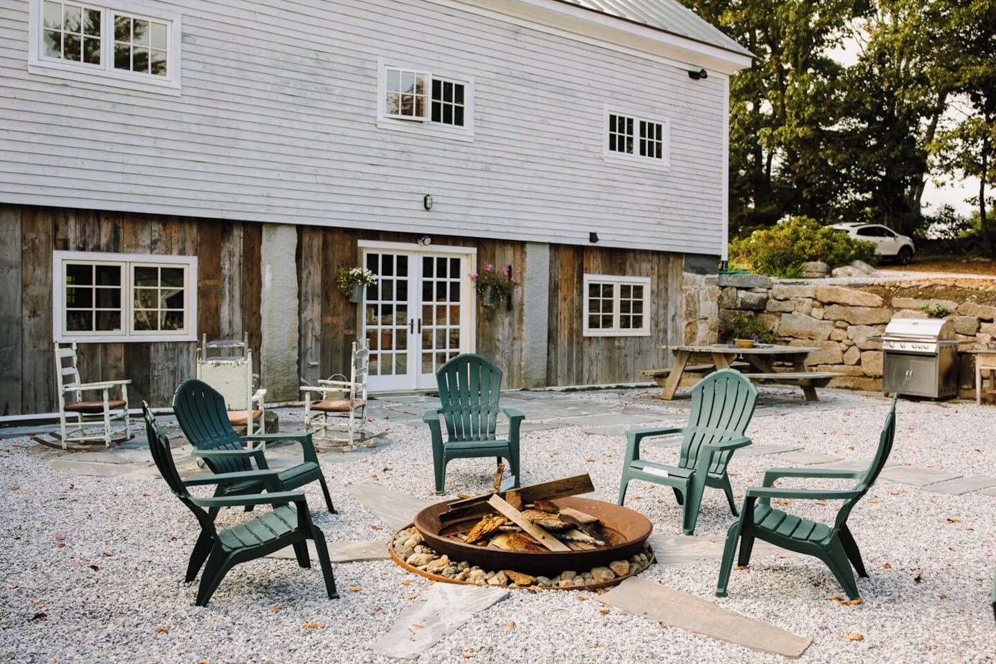 A rustic outdoor seating area with Adirondack chairs around a fire pit, a stone wall, and a wooden building with windows and a door.