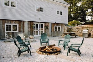 A rustic outdoor seating area with Adirondack chairs around a fire pit, a stone wall, and a wooden building with windows and a door.