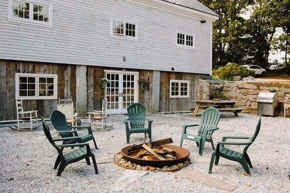 A rustic outdoor seating area with Adirondack chairs around a fire pit, a stone wall, and a wooden building with windows and a door.