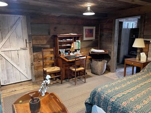 A rustic room with a wooden desk, a chair, a bed with patterned bedding, a lamp, and a framed picture on the wall.