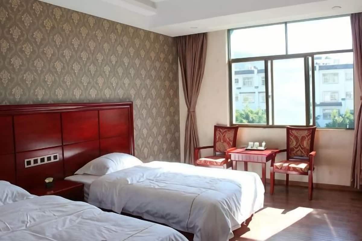Habitación de hotel con dos camas, cabecero de madera, una mesita, una silla y ventana con vista a edificios.