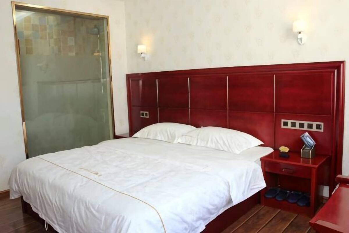 Una habitación de hotel con una cama grande, una mesita de noche, un cabecero rojo y una lámpara fijada a la pared.
