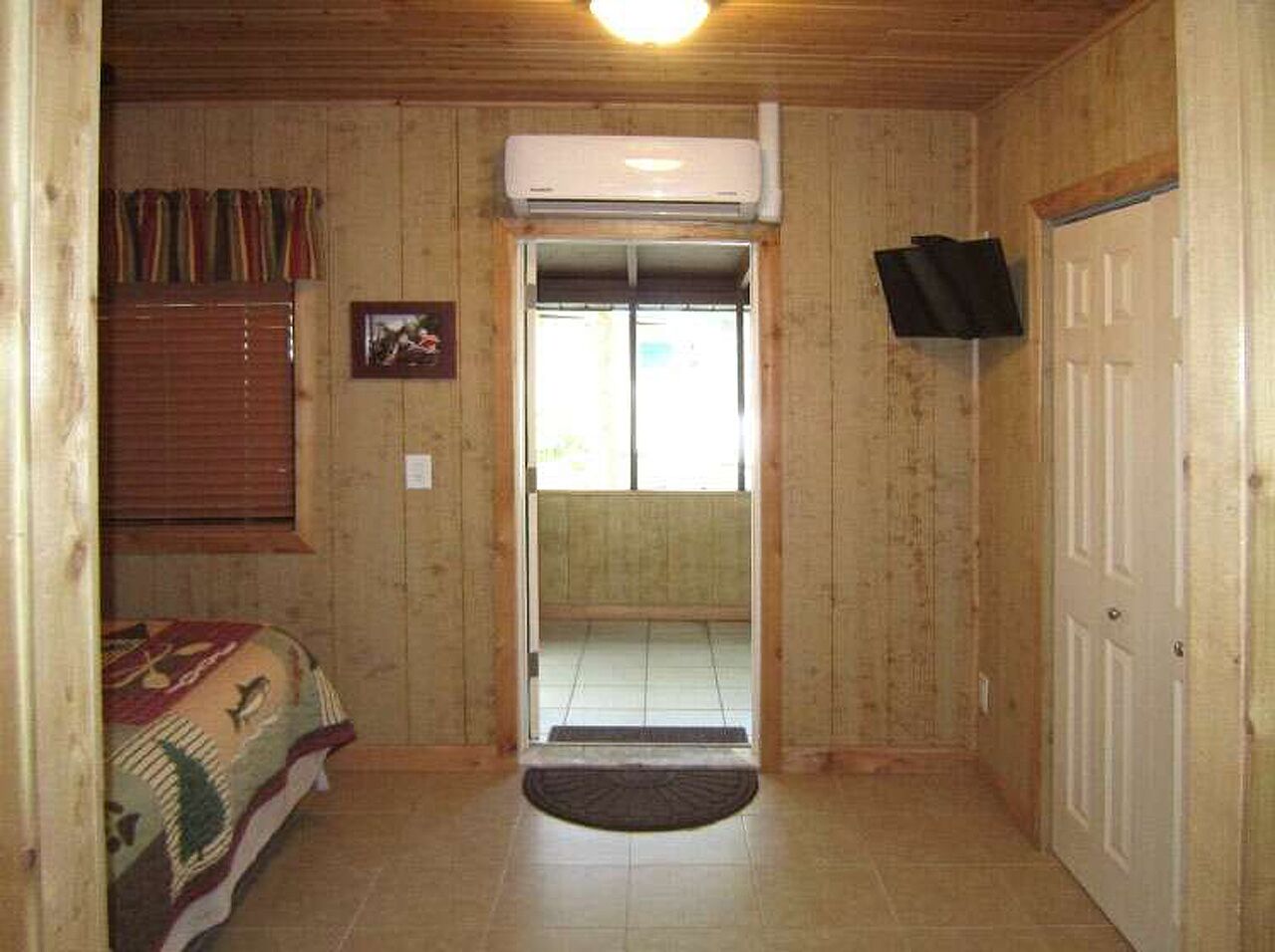 Ein Zimmer mit Holzwänden, einem Bett mit gemustertem Überwurf, einer Tür und einem an der Wand befestigten Fernseher.