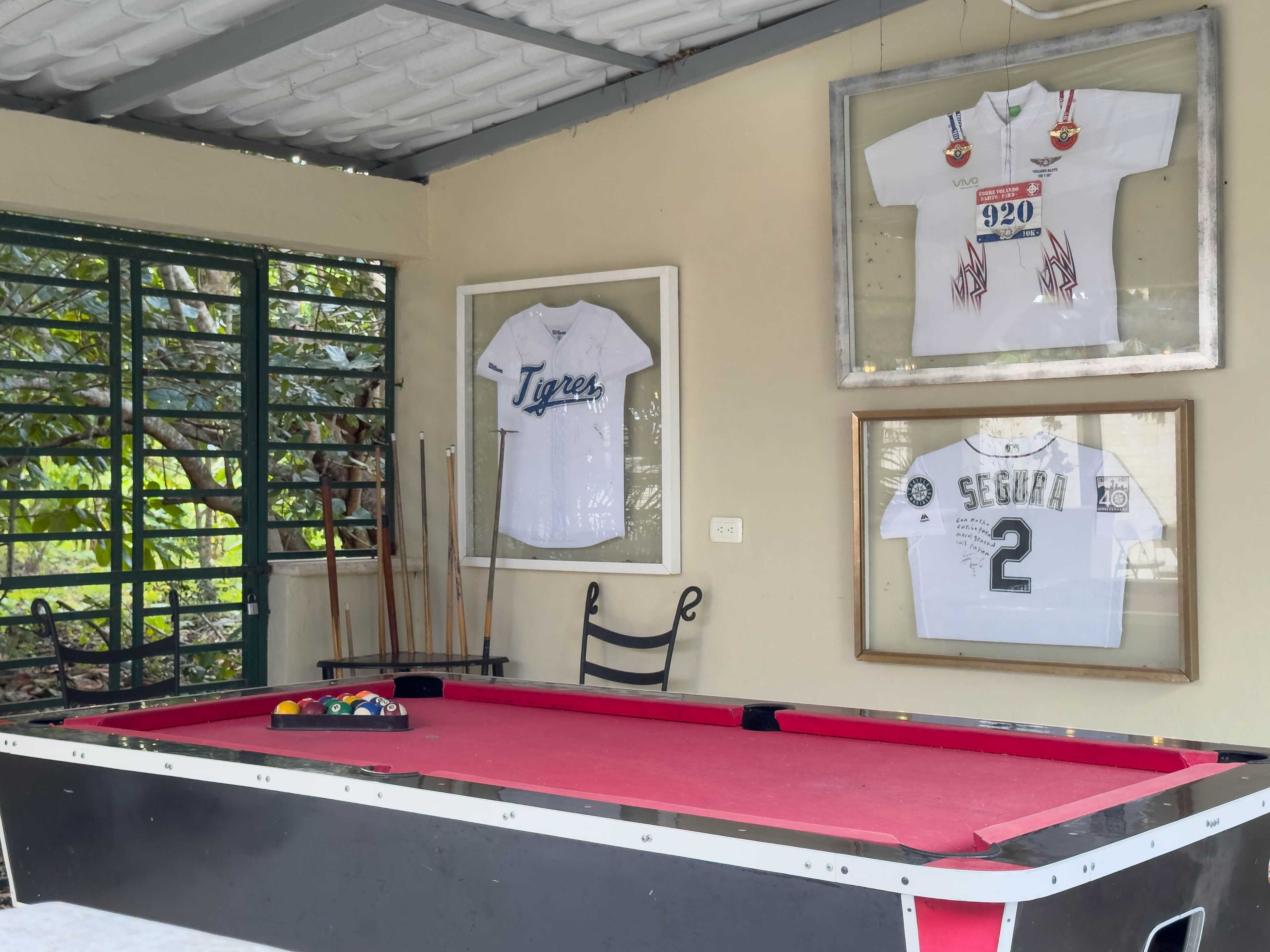 Una mesa de billar con superficie de fieltro rojo, rodeada de camisetas deportivas enmarcadas en la pared.