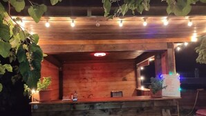 A wooden outdoor seating area with string lights, potted plants, and a signboard.