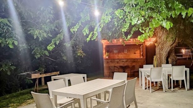 An outdoor dining area with white tables and chairs, a wooden barbecue grill, and a large tree providing shade.