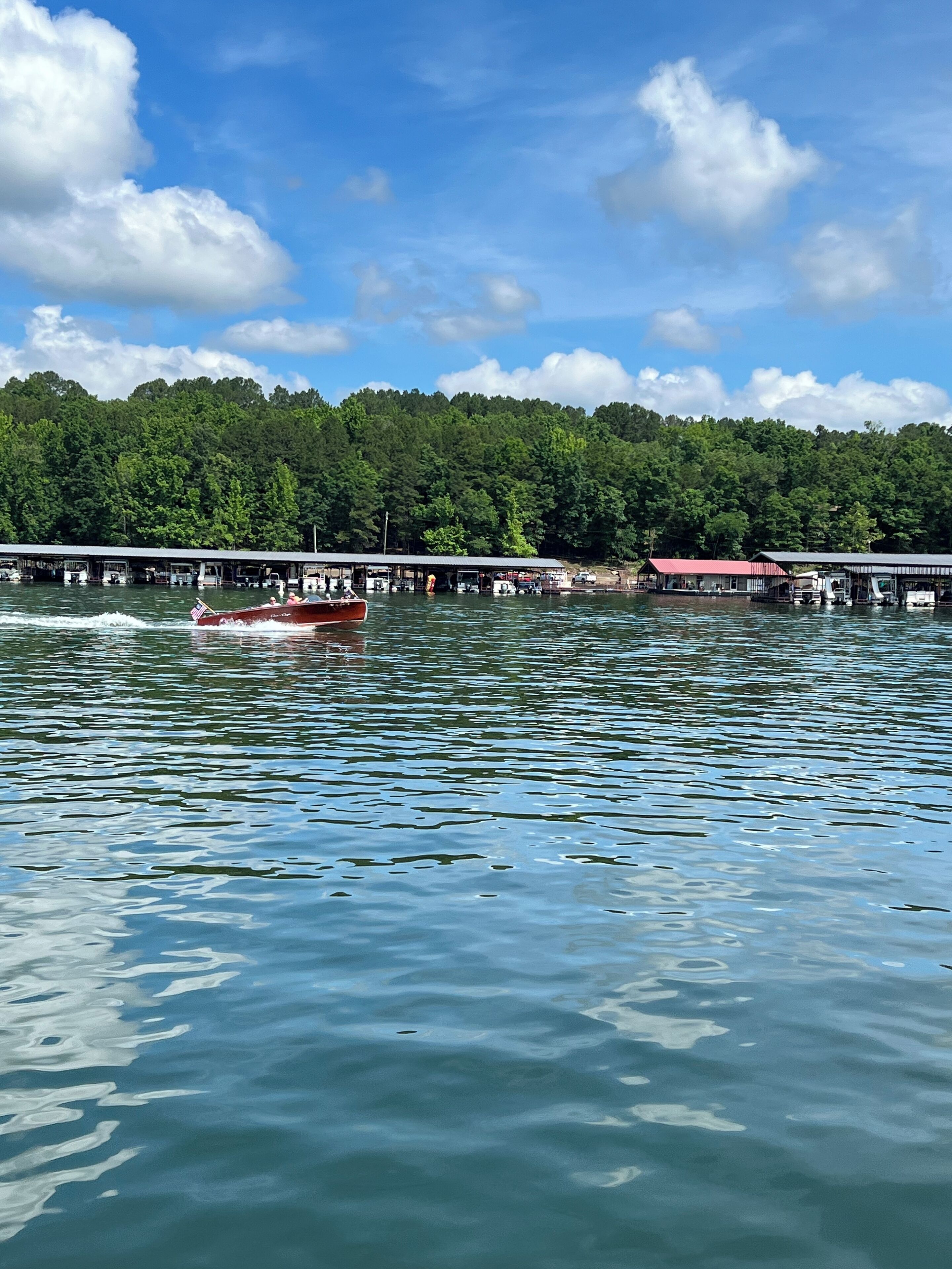 A boat on a lake with docked boats and a forested hillside in the background.